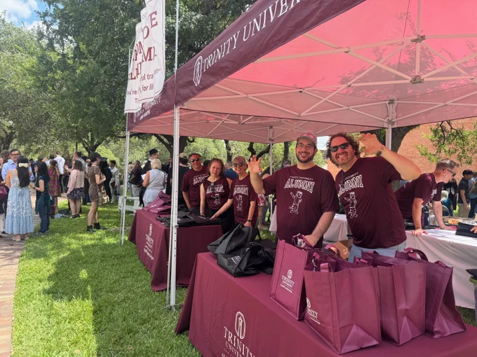 Alumni volunteers stand under a maroon Trinity University tent outside with maroon and black Trinity University bags on tables with maroon tablecloths.