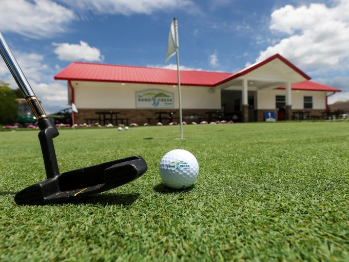 A golf putter and ball on a green near a hole with a flag. In the background, there's a building with a red roof featuring the Sand Creek Course at French Lick Resort logo. The sky is blue with scattered clouds.