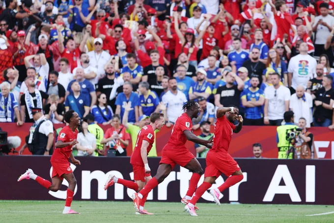 Cyle Larin celebrates scoring for Canada against Bosnia in the World Cup, with his eyes closed and both fingers on his ears as teammates chase him