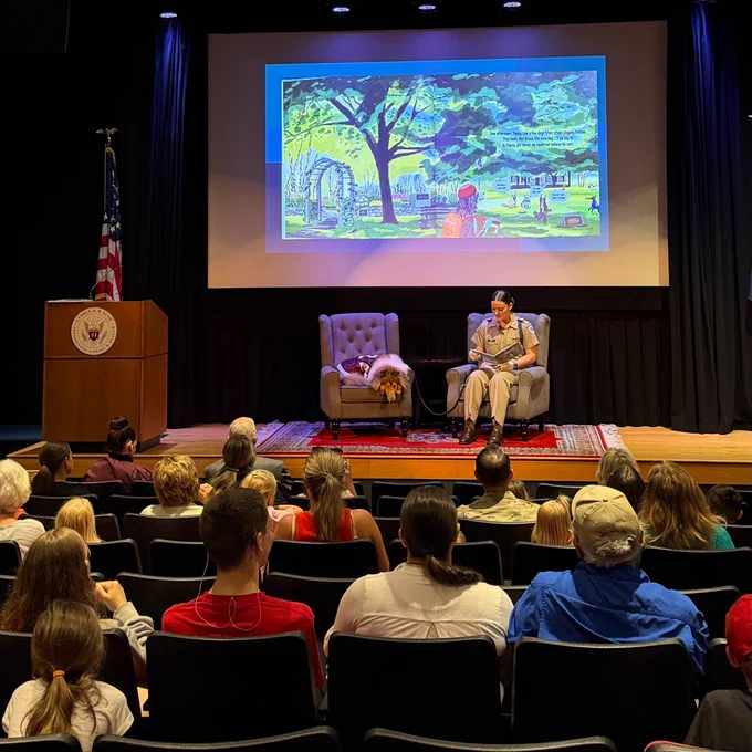 A person is seated on a stage, holding a book, in front of an audience. Beside them, a dog is resting on a chair. There is a colorful projection of a nature scene on a screen behind them. An American flag and a podium with a seal are visible on the left side of the stage.