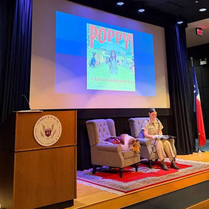 An individual in a beige uniform sits on a chair next to a dog on a stage. They are in front of a large screen displaying the book cover "POPPY! A Tale of Leading with Love" at the George H.W. Bush Presidential Library event space. There is a lectern with the library's seal and a Texas flag nearby.