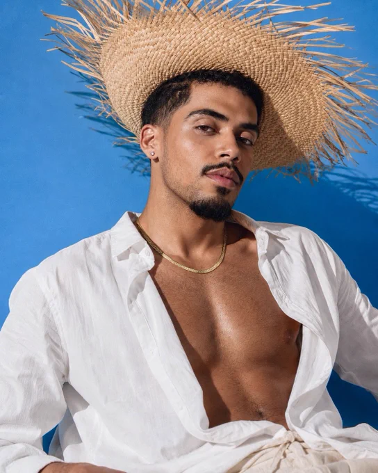 Matthew J. Sánchez, Founder & Perfumer of MATTEO PARFUMS, seen in new editorial campaign shoot for his fine fragrance brand. He’s wearing a decorative straw hat, white linen button up shirt, beige linen trousers, and wearing his gold jewelry.