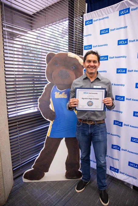 An individual dressed in business casual attire holds a certificate with "UCLA Staff Assembly" branding, recognizing Agustin Vega-Crespo as a scholarship winner. The person is standing next to a UCLA bear mascot cutout. The background includes a step-and-repeat banner with "UCLA Staff Assembly" logos and a window with blinds.