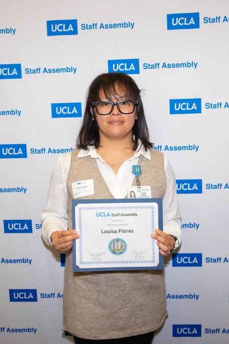An individual dressed in business casual attire and wearing a name badge holds a certificate that reads "UCLA Staff Assembly" with the name Louisa Flores as a scholarship winner. The background features a branded step-and-repeat banner with "UCLA Staff Assembly" logos.