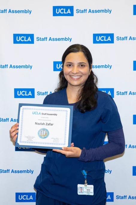 An individual wears blue scrubs and holds a certificate that reads “UCLA Staff Assembly” with the name Nazish Zafar as a scholarship winner. The background features a branded step-and-repeat banner with "UCLA Staff Assembly" logos.