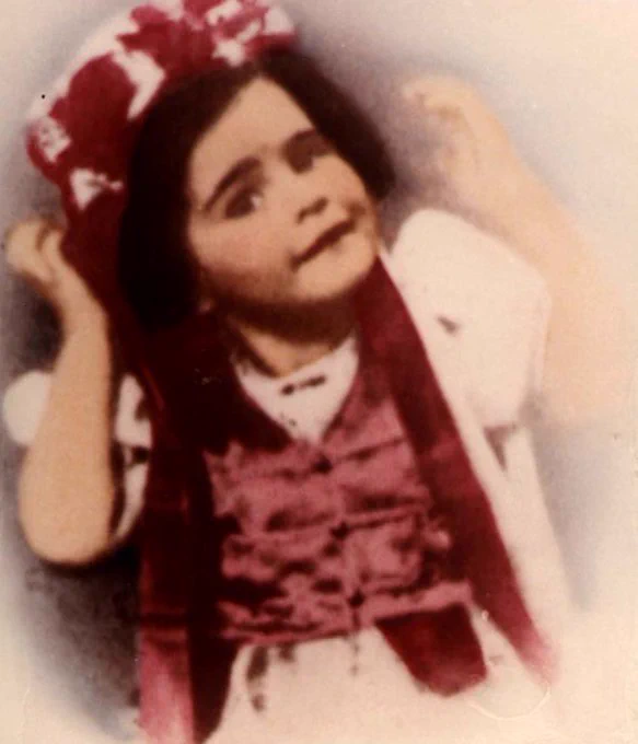 Color photograph of a young girl with a bow in their hair, wearing a vest and ribbons, holding their hands up in a playful manner.