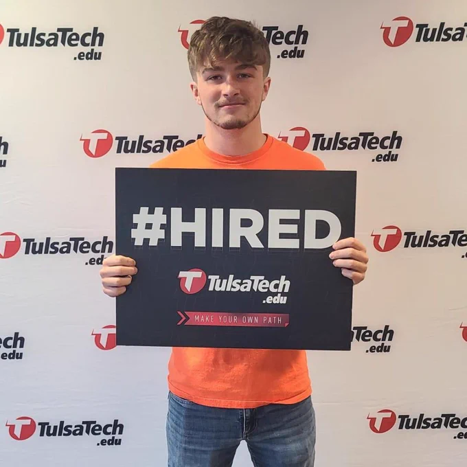 Person wearing an orange shirt holding a black sign that reads '#HIRED' with Tulsa Tech branding in the background.
