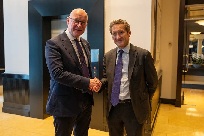 The First Minister and Carlyle Managing Director Richard Hoskins in business suits shaking hands in a modern office lobby with an elevator in the background.