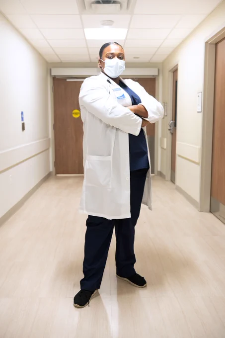 A masked healthcare worker wearing a lab coat over scrubs is standing with her arms crossed in a hallway.