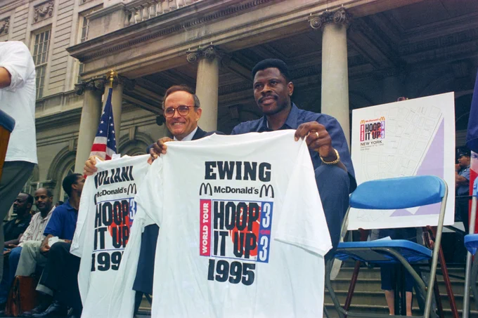 Two individuals seated outdoors hold up a white T-shirt featuring 'Ewing McDonald's Hoop It Up 3 on 3 World Tour 1995' text in front of a classical building with columns.