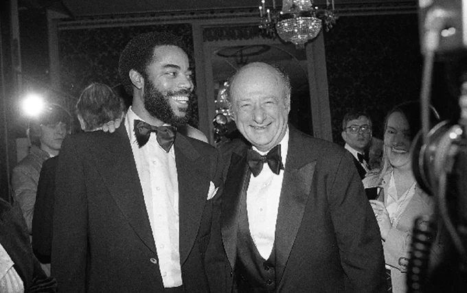 Two men dressed in tuxedos posing together at a formal event with a chandelier in the background.