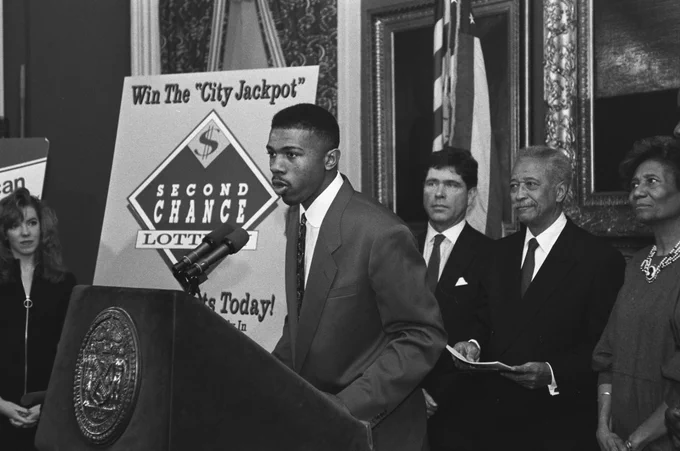 Man in suit speaking at podium with 'Second Chance Lottery' sign behind him in formal setting.