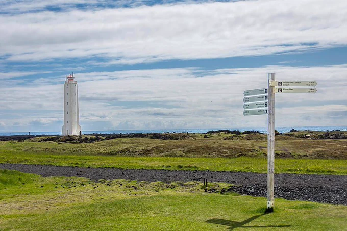 A tall, white lighthouse stands against a backdrop of expansive clouds. A signpost in the foreground directs travelers amid a green landscape. Photography by Debra Martz