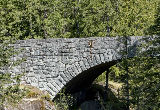 Stone arch bridge surrounded by green forest, with sunlight and shadow falling across the textured gray masonry.