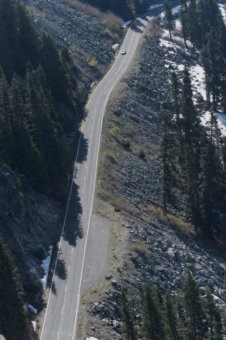 Curving mountain road climbing through a rocky alpine landscape, with steep slopes, evergreen trees, and bright sunlight creating strong shadows.