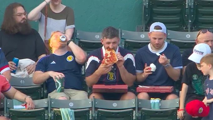 Three Scottish soccer fans are sitting in the stands at Fenway Park. One is chugging a beverage, one is eating pizza and one fan is eating ice cream.