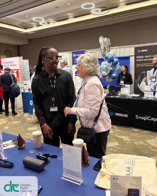 Photo 3: Tremmel Watson is candidly standing at a convention booth, trying out a demo glasses product. In front of him, he is interacting with an older woman with gray wavy hair and is wearing everyday glasses, a pink sweater, and purse.