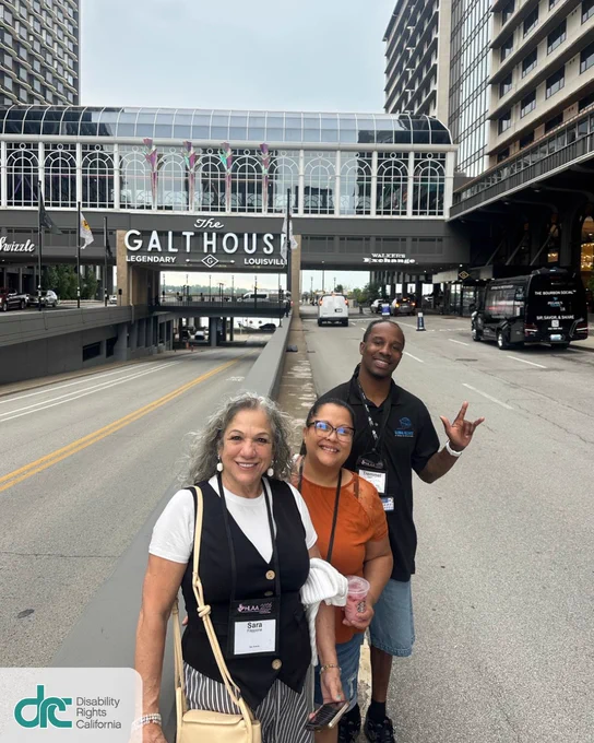 Photo 1: Three people smiling and posed together on an empty, large, 5-lane paved road between two large buildings and a windowed/mirrored bridge connecting the two. The bridged building has colorful flags and large signage: The Galhouse Legendary Louisville. Left is Sara, an older woman with long curly gray hair. Middle, a woman with dark hair pulled back. Right is Tremmel, a Black man with locs tied back. He holds up ASL for "I love you"