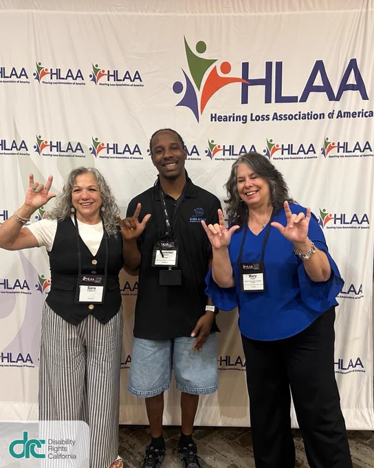 Photo 2: Sara, Tremmel, and Mary smiling and posed together, holding up the ASL sign for "I love you." They are standing in from of a photo back drop which has the logo for Hearing Loss Association of America (HLAA)