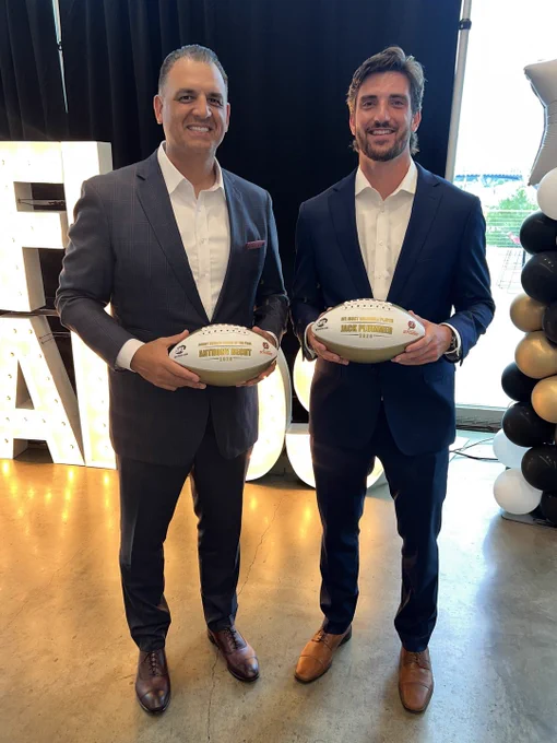 Two men in suits holding footballs at an awards event. They are smiling and standing in front of a lit sign and balloon decorations.