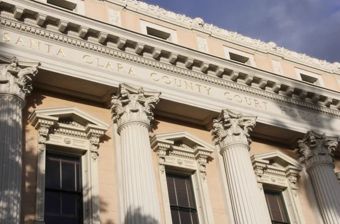 Exterior view of the historic Santa Clara County Courthouse, featuring tall Corinthian columns and ornate architectural details. The words “Santa Clara County Court House” are engraved across the upper facade beneath a clear blue sky.