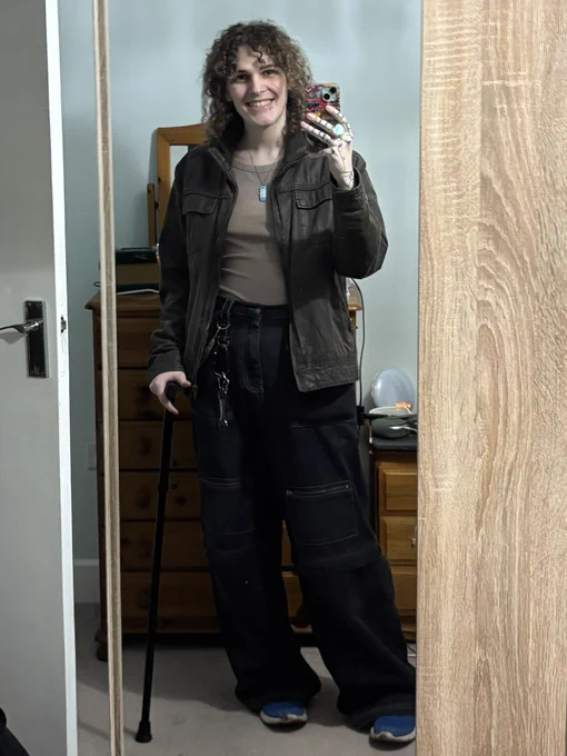 A picture of me taking a mirror selfie in a brown jacket, brown vest and black trip pants, holding a black cane.