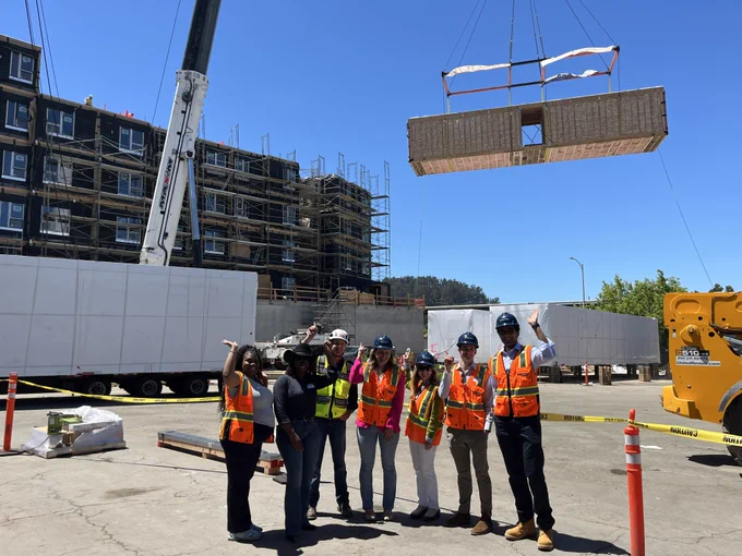 Assemblymember Wicks and a group of others wearing high visibility vests and hard hats stand in front of the active construction site of El Cerrito Plaza while a crane hoists a volumetric modular unit through the sky behind them towards the upper levels of the new building. 