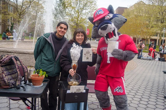 A person in a green jacket and another with a dark top are smiling next to a mascot dressed in a red outfit with "Stony Brook" on it. They are standing by a small table with a potted plant and a contraption holding a tall glass. A fountain and trees are visible in the background.