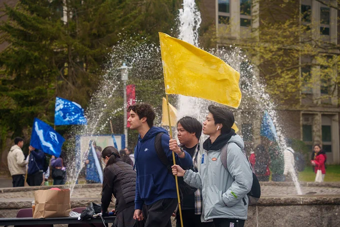 A group of people stands in front of a large, active fountain, holding a yellow flag. Blue flags are also visible in the background. There is a bustling environment with several individuals gathered around. The setting appears to be an outdoor area with trees and a building in the background.