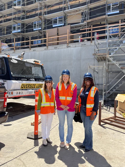 Assemblymember Wicks and two other women all wearing high visibility vests and hard hats stand together in front of the new building of the El Cerrito Plaza project being constructed with volumetric modular components. 