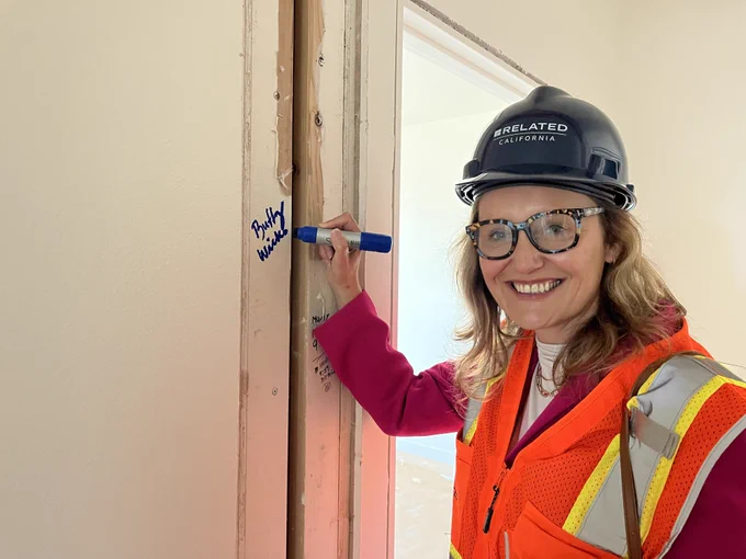 Assemblymember Wicks smiles and signs her name to the drywall inside one of the units constructed with prefabricated modules at the El Cerrito Plaza project site while wearing a high visibility vest and hard hat. 