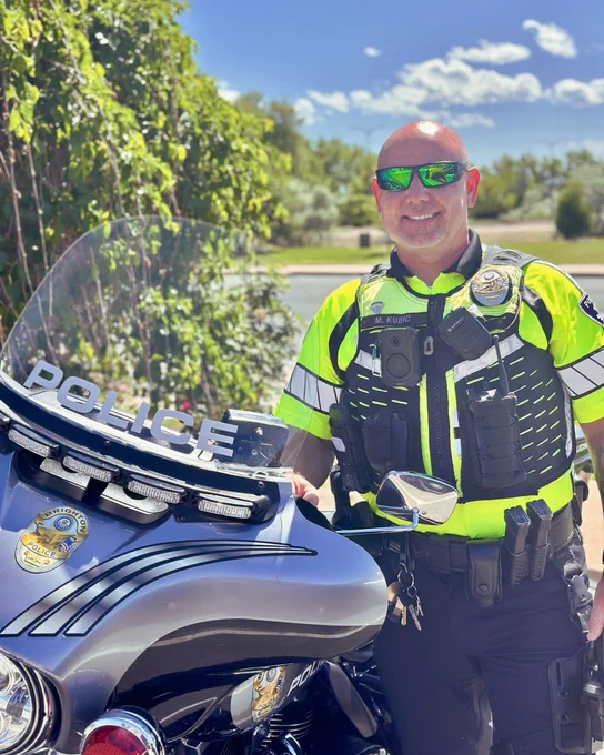 A police officer in a bright yellow and black uniform stands next to a marked police motorcycle with a clear windshield displaying the word 'POLICE,' set against a sunny outdoor background with greenery and a blue sky.