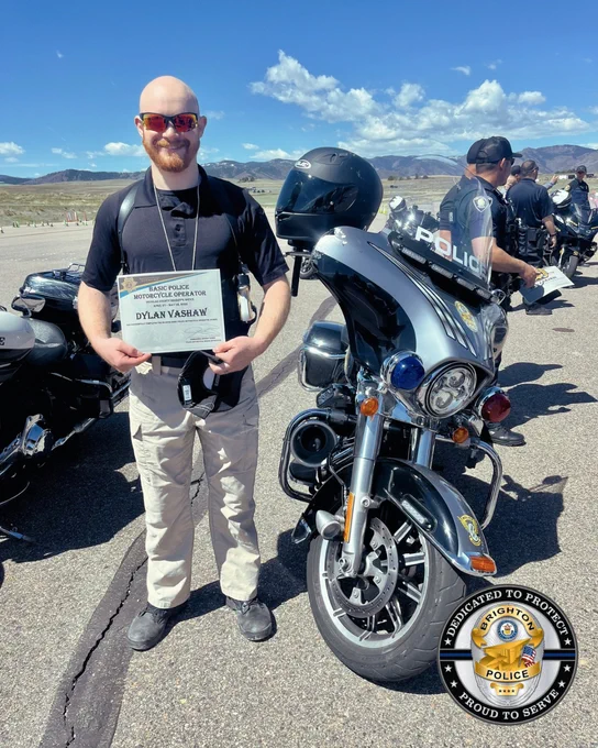 A person stands beside a police motorcycle on a sunny day, holding a certificate that reads 'Basic Police Motorcycle Operation Dylan Vaslaw,' with other officers and motorcycles lined up in the background under a clear blue sky and mountainous landscape.