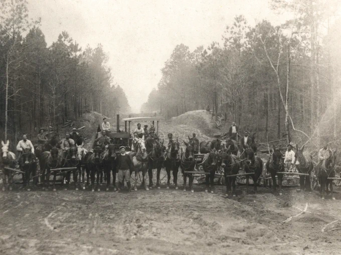 A historical black and white photo depicts a group of individuals posed with multiple horse- and mule-drawn plows on a dirt road in a forested area. Trees line both sides of the path, with bare branches visible. The group appears to be part of a road crew.