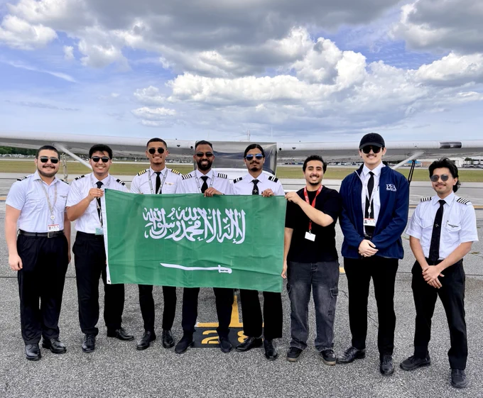 A group of people standing on a tarmac, holding a large flag of Saudi Arabia. They are dressed in aviation uniforms, and there is an airplane in the background under a partly cloudy sky.