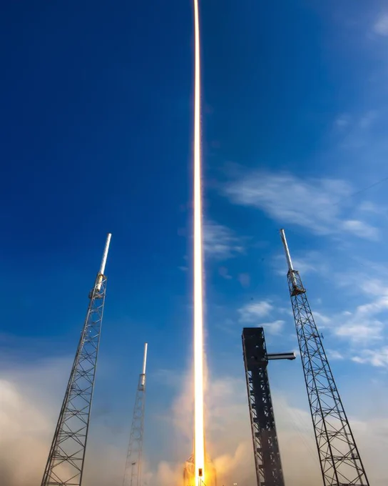 A SpaceX rocket is launching into a clear blue sky. The bright trail of the rocket's ascent is visible between several tall launch towers.