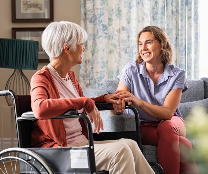 Two people sitting in a living room holding hands and having a conversation. One person is sitting in a wheel chair. 