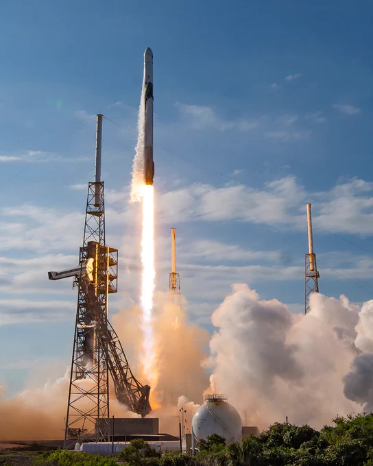 A SpaceX rocket is launching into a blue sky, leaving a trail of smoke and bright flames. It is surrounded by metal launch structures, and white clouds of smoke billow at the base.