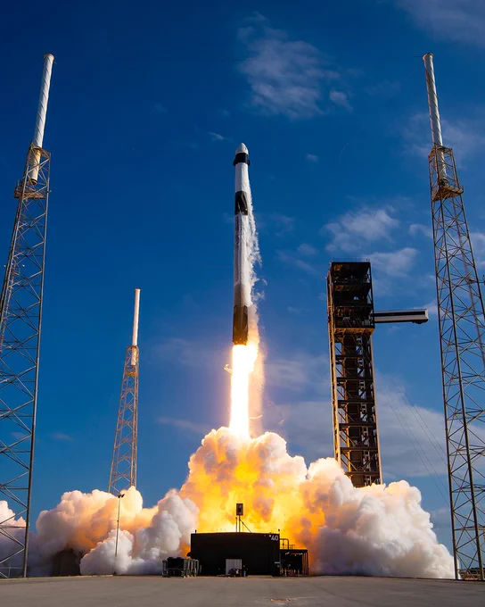A SpaceX rocket is launching into a clear blue sky, surrounded by smoke and flames. It is vertically ascending from a launch pad supported by tall structures.