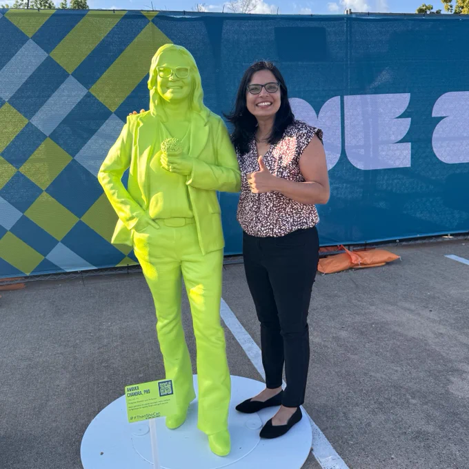 A person stands next to a bright yellow-green statue, smiling and giving a thumbs-up. They are outdoors, with a blue and green patterned backdrop behind them.