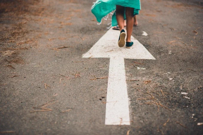 A child walks on a paved road with a large white arrow pointing forward. The person is wearing a teal garment and dark shoes. Pine needles are scattered on the road.