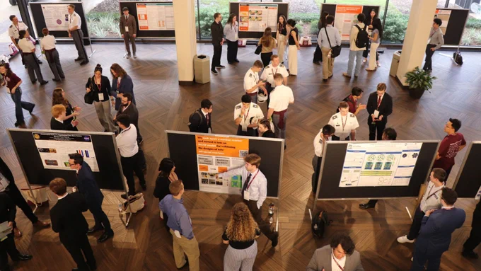 A group of people are gathered in a spacious room with several poster boards on display. The setting appears to be a professional or academic event, such as a conference or exhibition. Attendees are standing in small groups, engaging in discussions, and some are viewing the posters. The room features large windows in the background, allowing natural light to illuminate the hardwood floor.