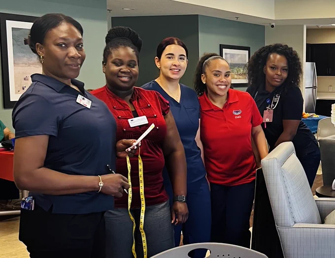 Group of five nursing students standing indoors, some wearing name tags and holding measuring tools, in a casual professional setting with framed artwork on the walls.