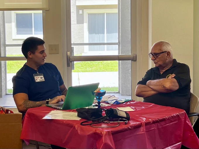 A healthcare worker with a laptop consults an elderly man seated across a table covered with a red cloth in a bright room.