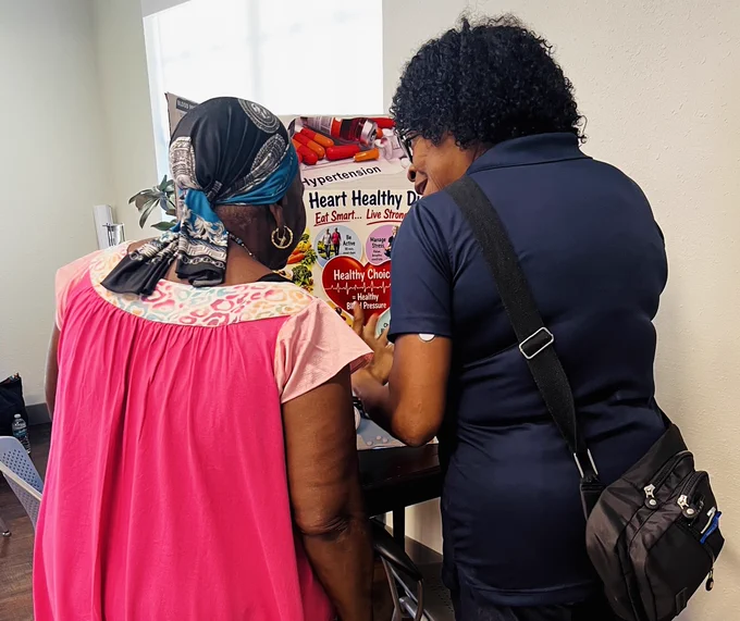 Two women stand together reviewing a colorful health poster about hypertension and heart health in a well-lit room.
