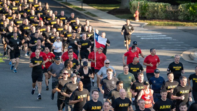 A U.S. Army Soldier in black physical training gear turns toward the camera and gives a thumbs-up while participating in the Army's 251st Birthday Run at Joint Base Myer-Henderson Hall, Virginia, June 12, 2026. He is running alongside a large formation of Soldiers, service members, civilians, and international partners.