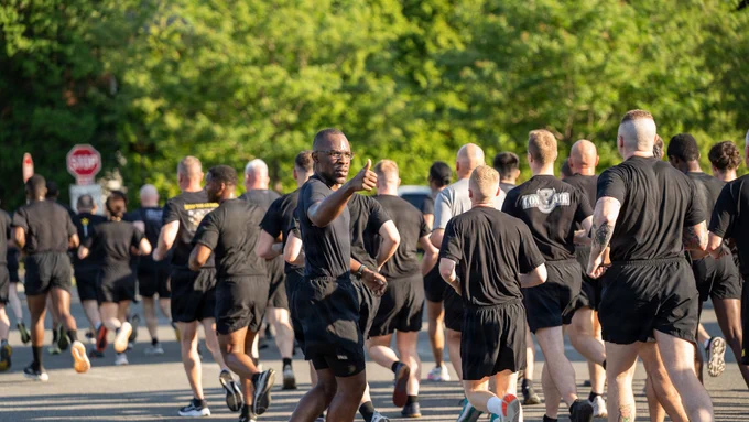 A U.S. Army Soldier in black physical training gear turns toward the camera and gives a thumbs-up while participating in the Army's 251st Birthday Run at Joint Base Myer-Henderson Hall, Virginia, June 12, 2026. He is running alongside a large formation of Soldiers, service members, civilians, and international partners.