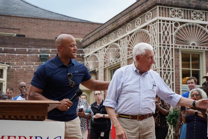 A person is speaking at a podium with a banner that has partially visible lettering. They are holding a microphone and gently touching the shoulder of another person who is walking past. The scene takes place outdoors with a decorative brick and wrought-iron structure in the background, surrounded by a small crowd.