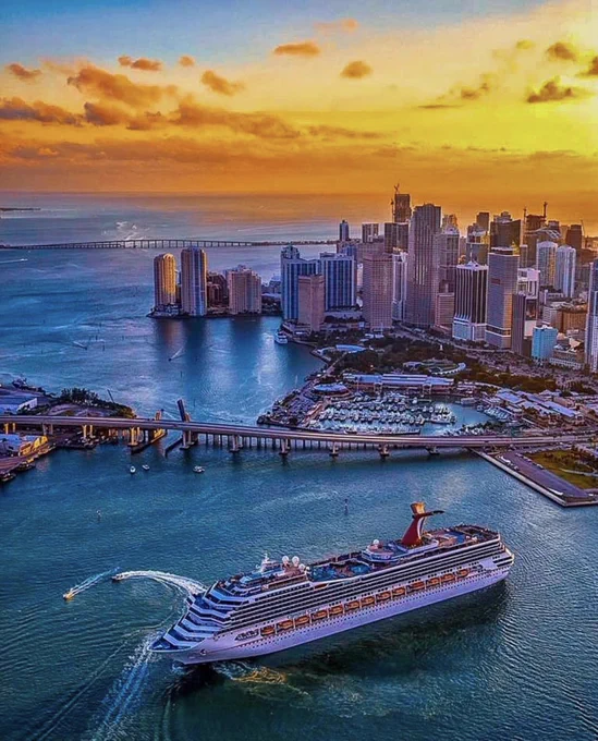 An aerial view shows a large cruise ship with distinctive red and blue funnels, sailing on blue water. Behind it, a vibrant city skyline rises under an orange and yellow sunset. Bridges span the water, and small boats are visible near a marina.