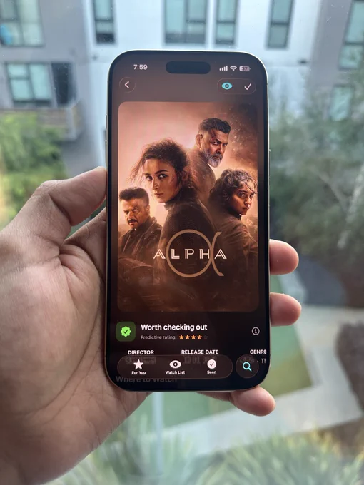 Phone displaying a movie poster for "Alpha" with four characters, glowing orange background. Text underneath says “Worth checking out” with star ratings. Hand is holding the phone against a blurred outdoor background.